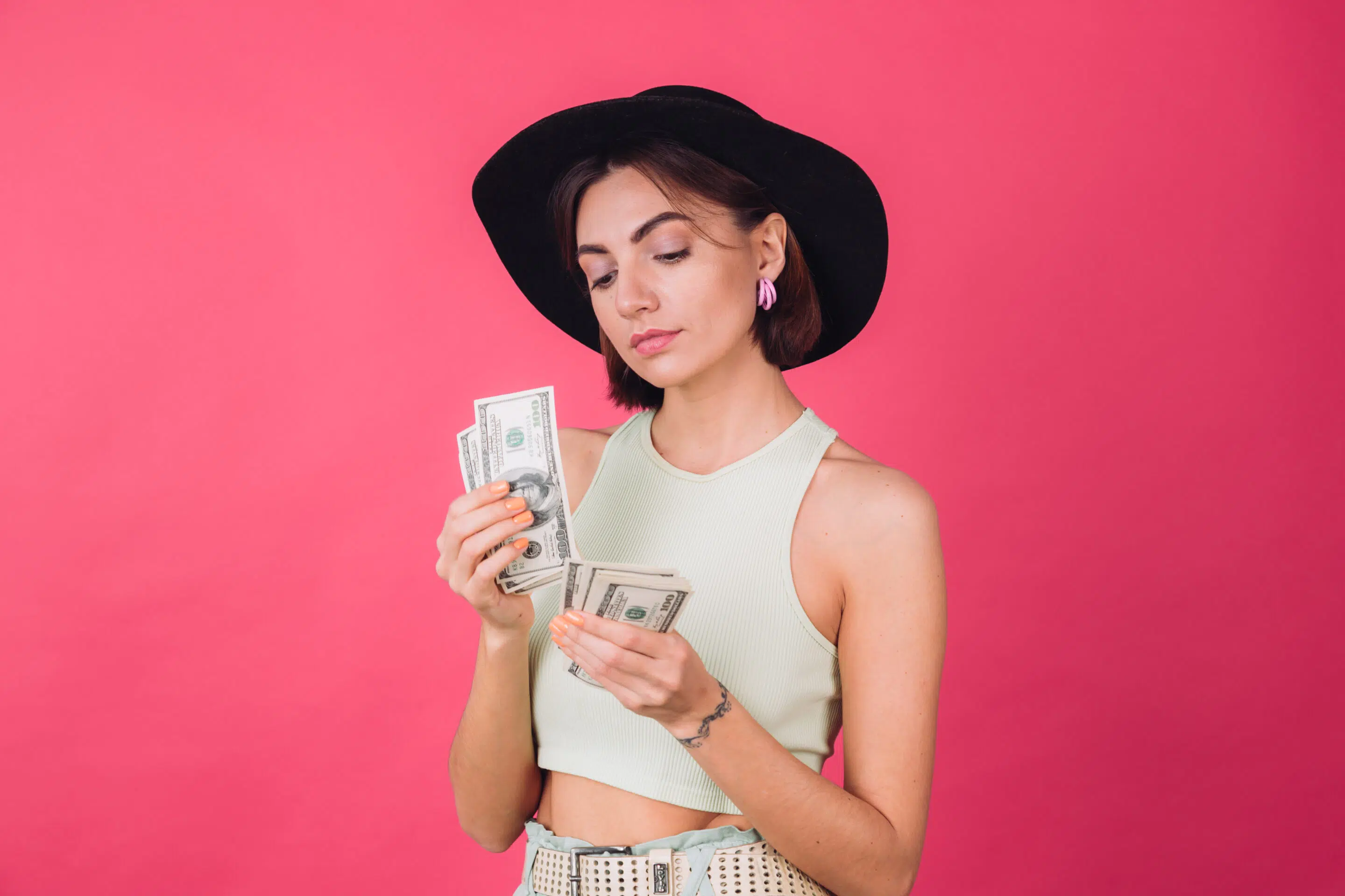 Stylish woman in hat on pink red background isolated space smile excited hold fan of 100 dollars money thoughtful count money